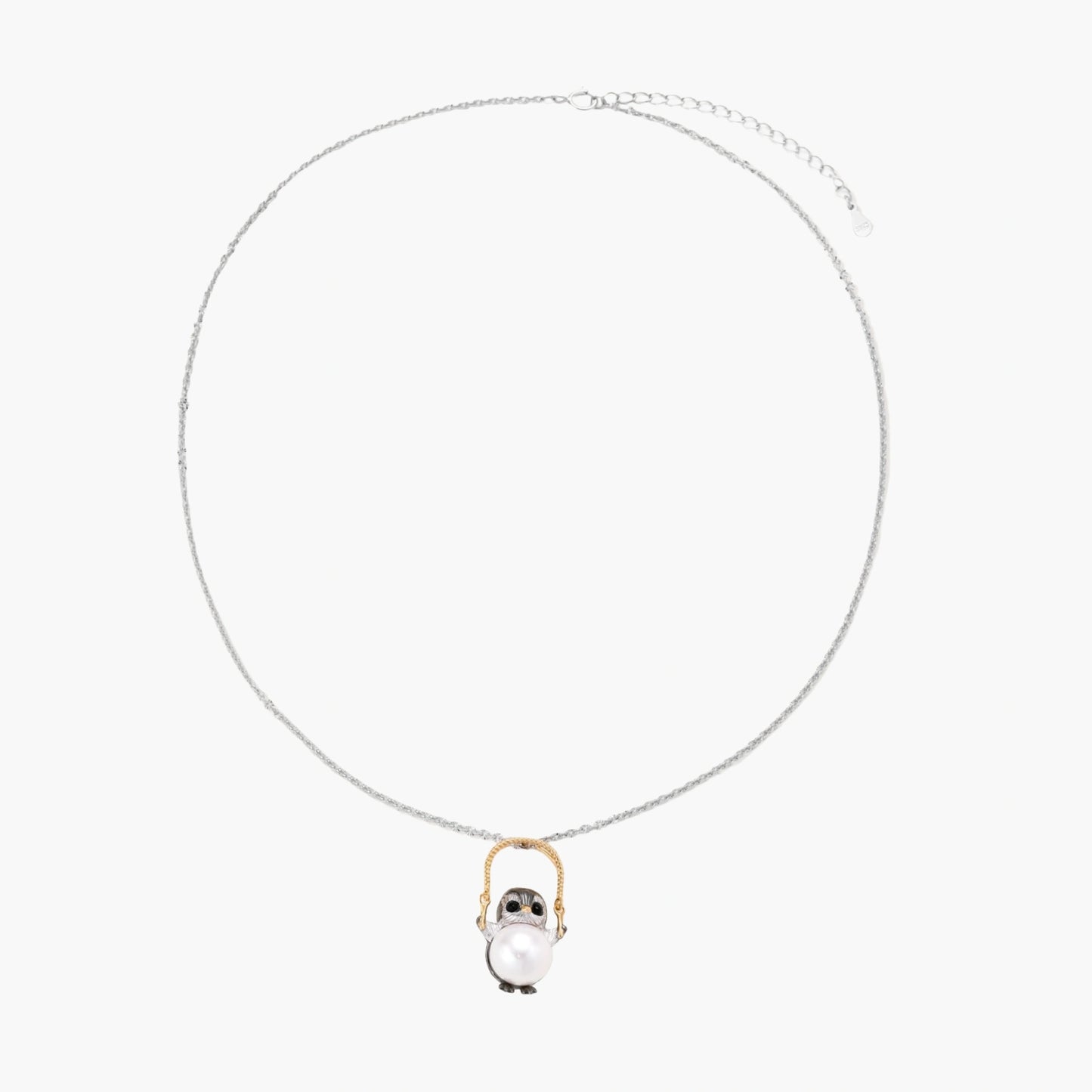 Whimsical owl pendant holding a white pearl on a gold swing, rhodium-plated silver chain on cream background.